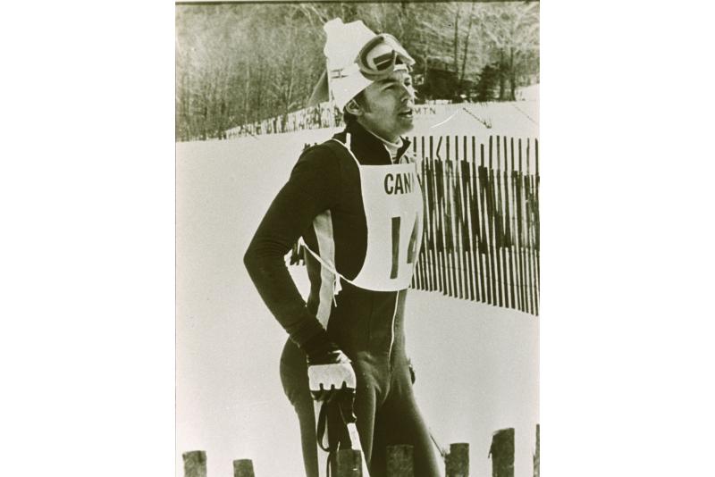 On a mountainside, Otto stands with ski poles and a racing suit. 